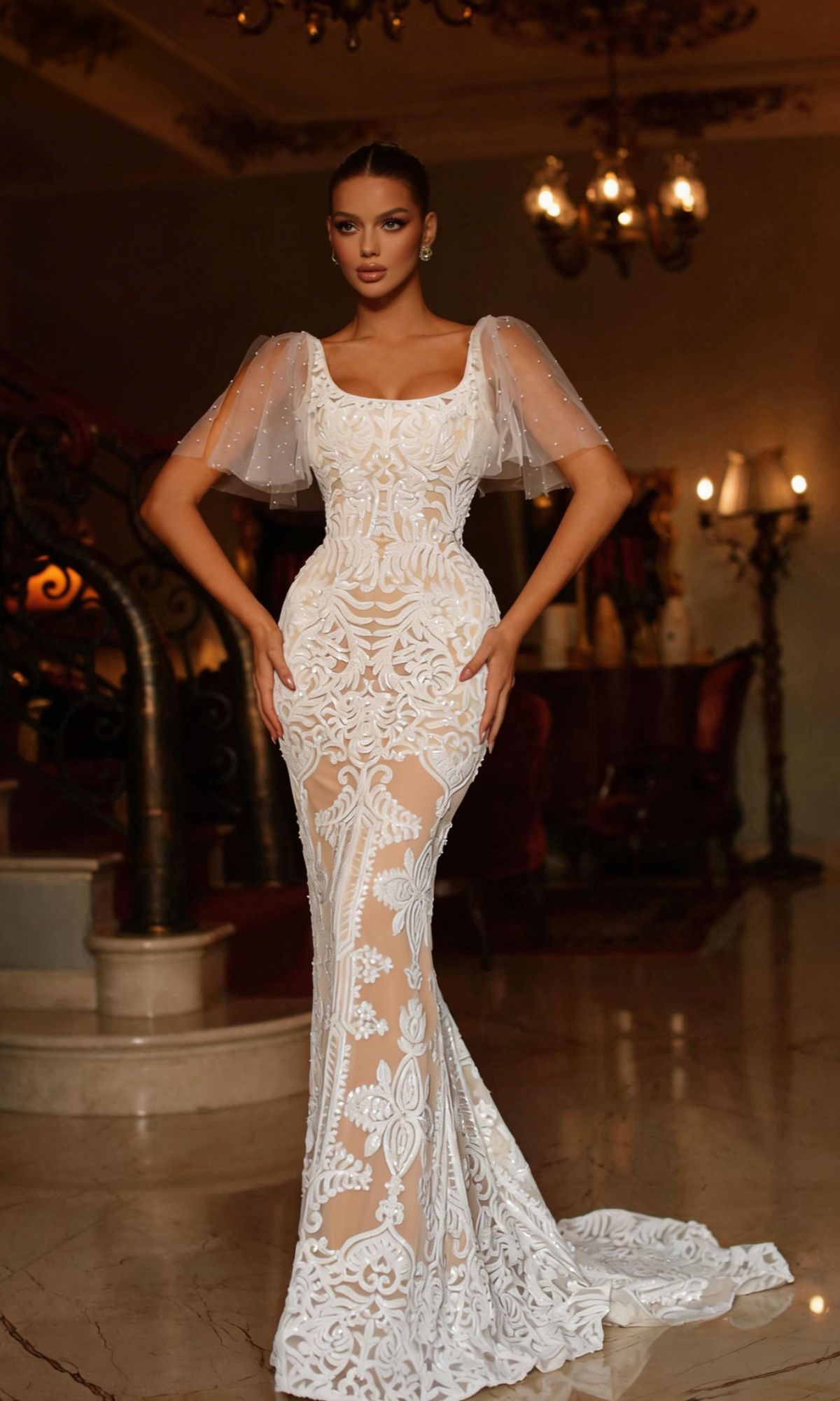 Woman wears the Tina Holly TW111 short sleeve white formal gown, featuring bead-embellished sheer sleeves and lace details, while standing indoors on a polished marble floor.